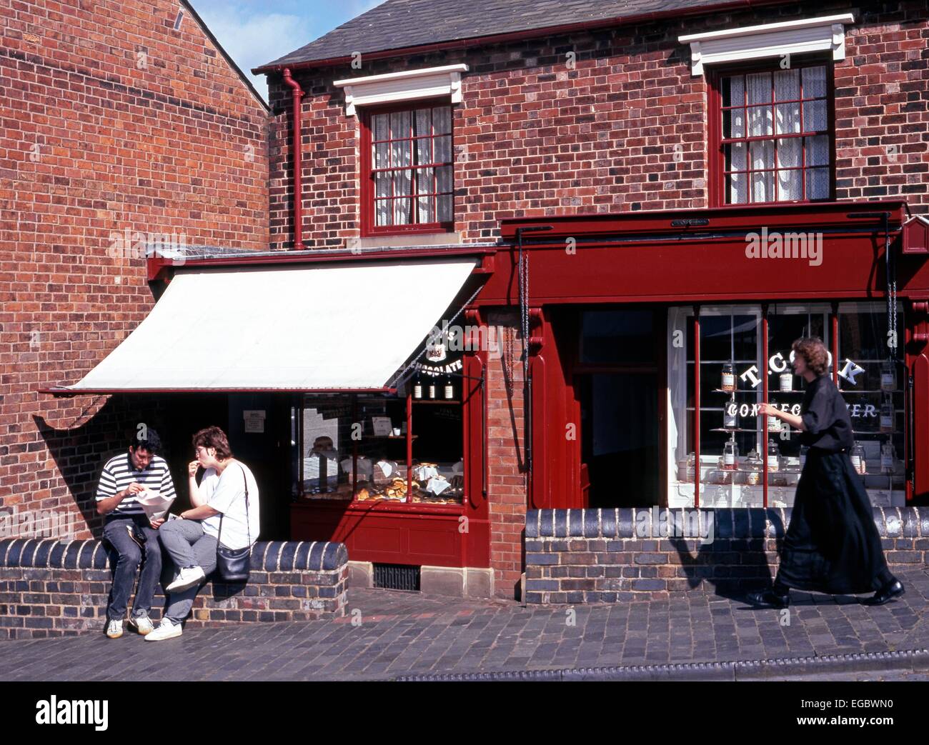 Couple sitting eating on a wall outside a Victorian shop with a Victorian woman walking along in the Black Country Living Museum Stock Photo