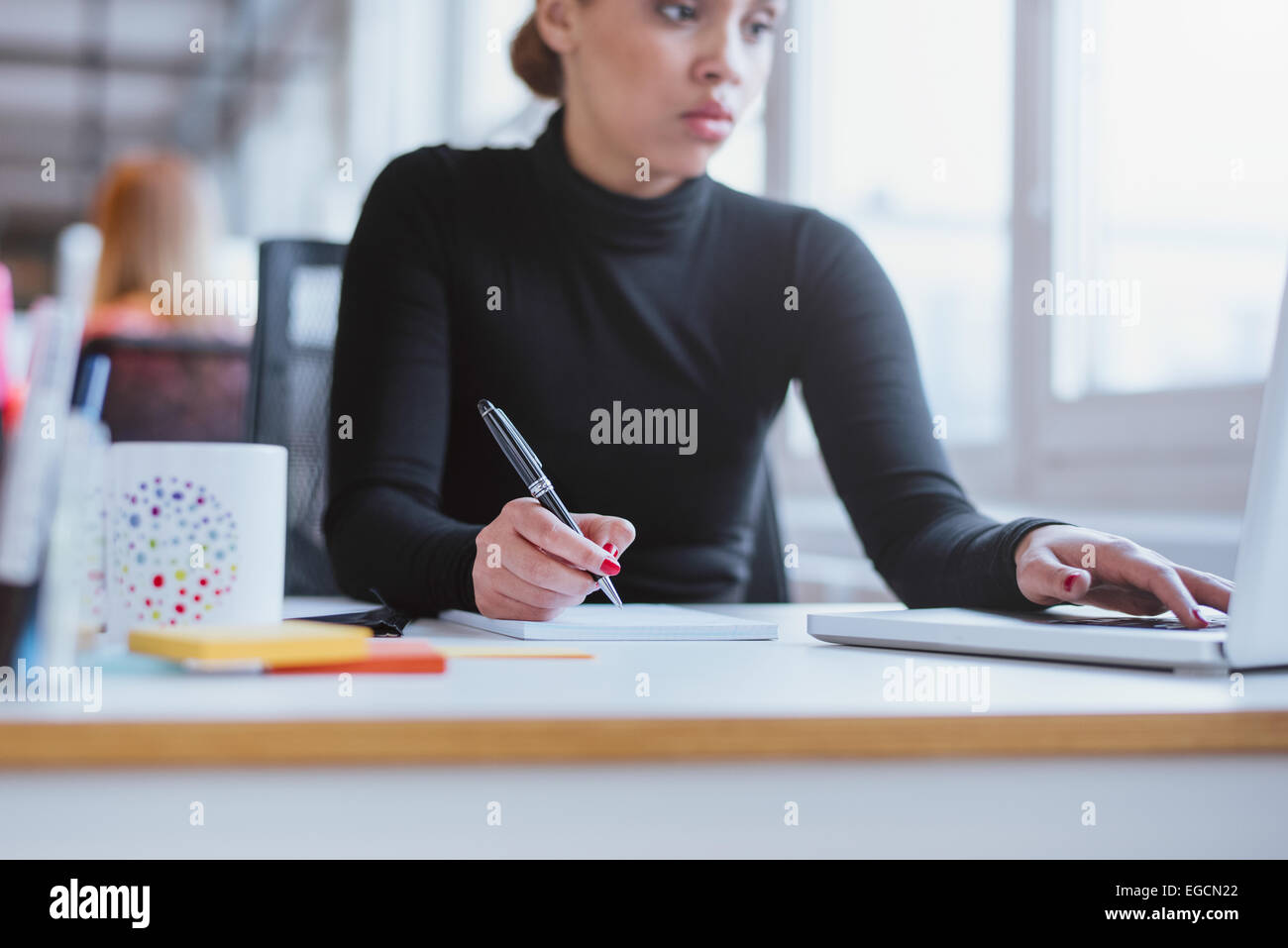 Young woman taking notes from laptop. Female executive working her desk using laptop and writing notes. Stock Photo
