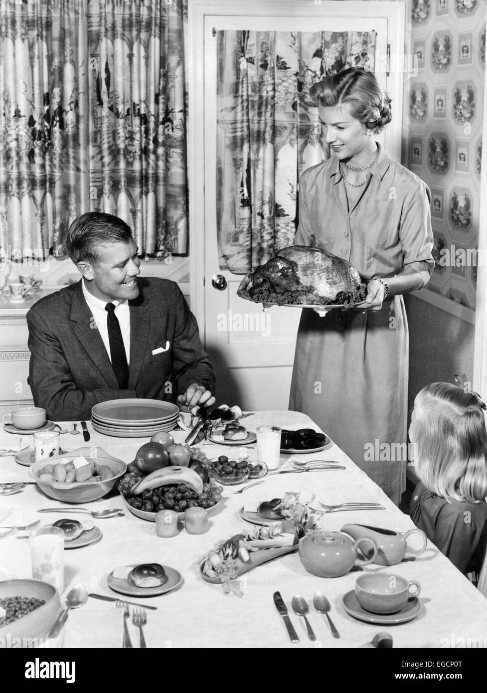 1950s THANKSGIVING FAMILY AT DINING ROOM TABLE MOM HOLDING TURKEY ON PLATTER PRINT CURTAINS TABLE SETTING WALLPAPER Stock Photo