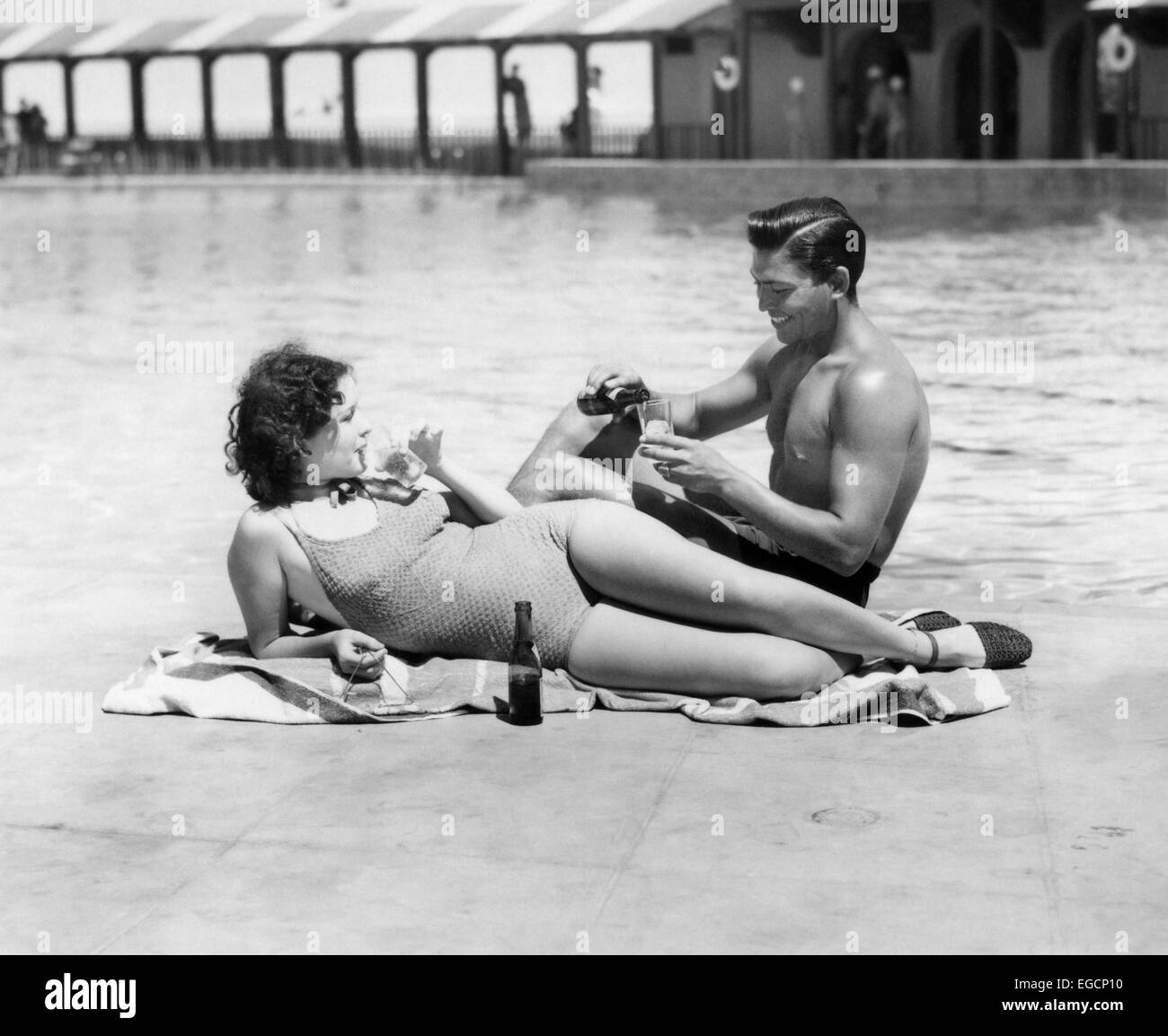 1930s 1940s COUPLE WEARING BATHING SUITS SITTING  RELAXING POOL SIDE DRINKING BEER Stock Photo
