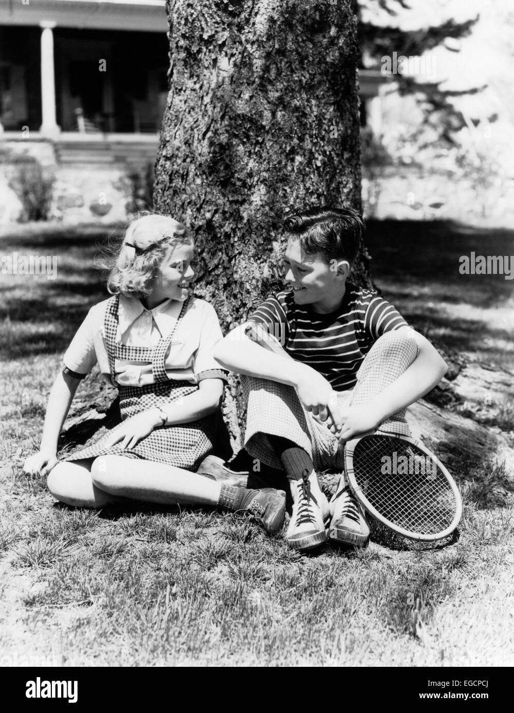 1930s 1940s BOY AND BLOND GIRL SITTING OUTSIDE BY TREE TRUNK TALKING HOLDING TENNIS RACKET Stock Photo