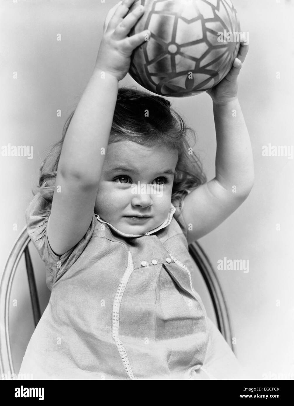 1930s 1940s LITTLE GIRL TODDLER HOLDING TOY BALL ABOVE HER HEAD WITH BOTH ARMS Stock Photo