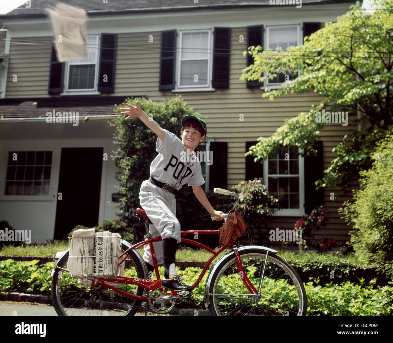1960s BOY IN BASEBALL UNIFORM RIDING BICYCLE DELIVERING NEWSPAPERS Stock Photo