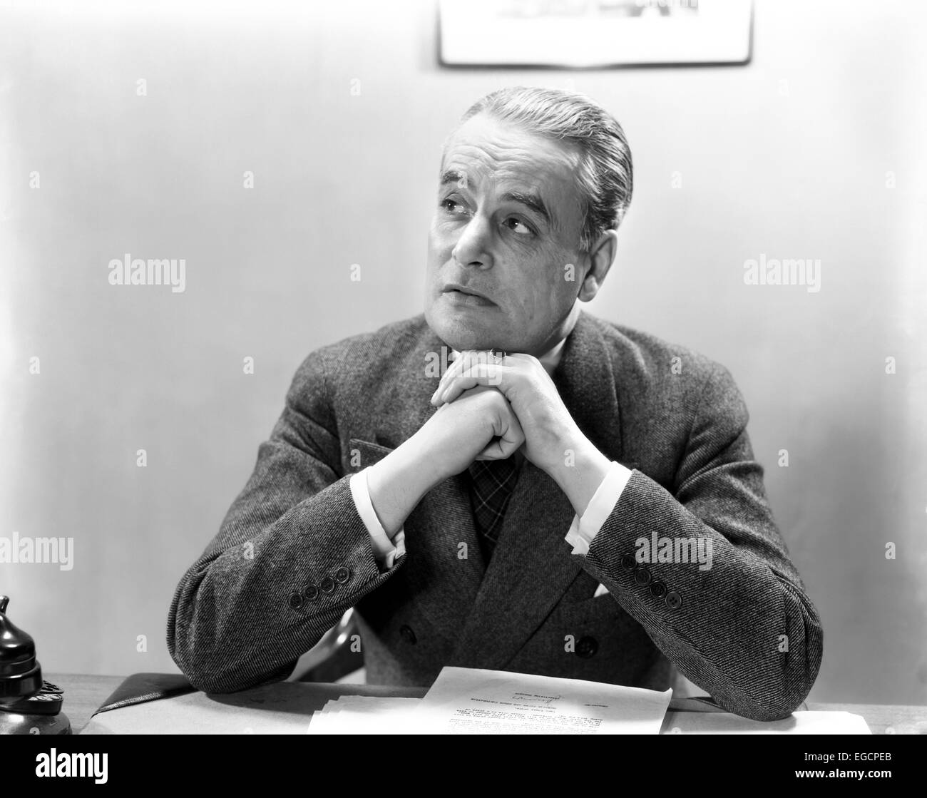 1930s 1940s MAN PORTRAIT AT DESK BUSINESSMAN WEARING SUIT HANDS CLASPED UNDER CHIN LOOKING OFF TO SIDE Stock Photo