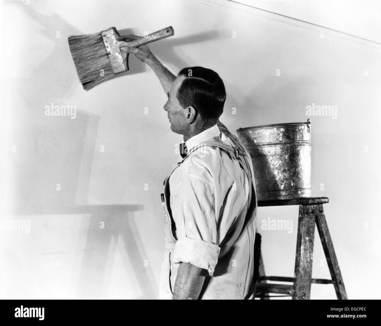 1930s 1940s MAN IN WHITE OVERALLS BY STEP LADDER ZINC PAIL BUCKET APPLYING PAINT GLUE WITH LARGE BRUSH TO PREPARE FOR WALLPAPER Stock Photo