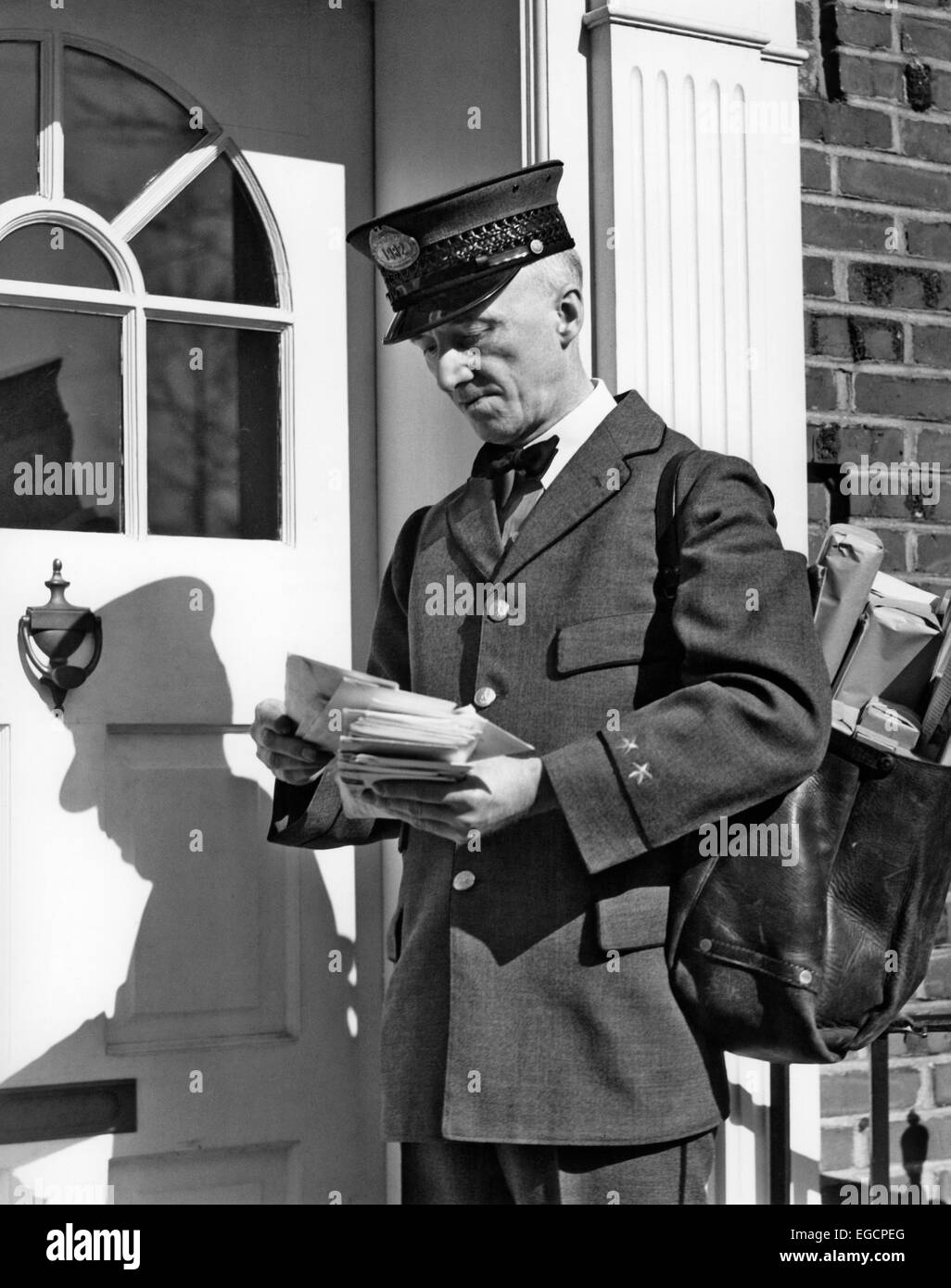 1930s 1940s POSTAL SERVICE UNIFORMED MAILMAN DELIVERING MAIL STANDING ON DOORSTEP SORTING LETTERS Stock Photo