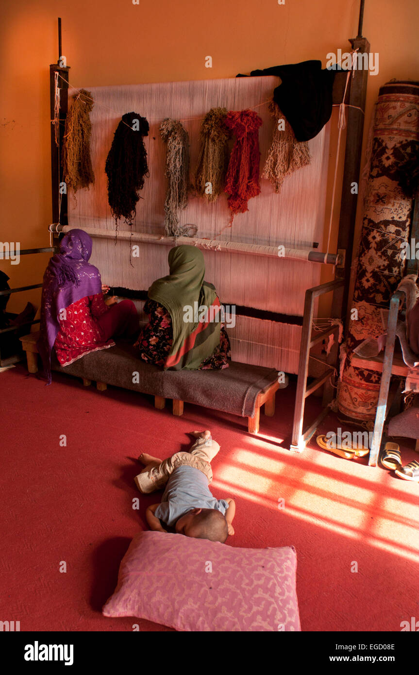 Herat Women's prison - weaving workshop - 2 women weave while a baby lies on the carpet Stock Photo