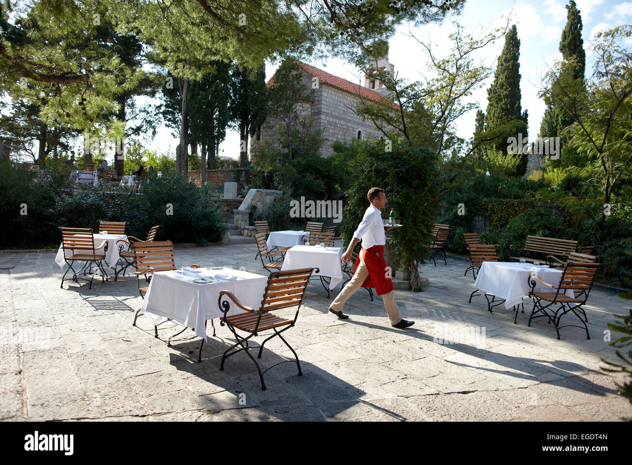 Waiter and coffee tables on the Piazza under trees, Aman Sveti Stefan, Sveti Stefan, Montenegro Stock Photo