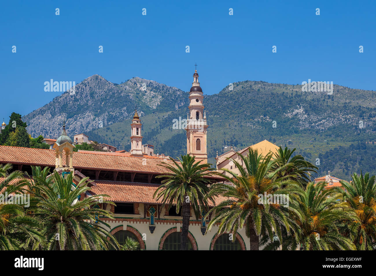 Catholic church bell tower among houses and palms and mountain on background under blue sky in Menton, France. Stock Photo