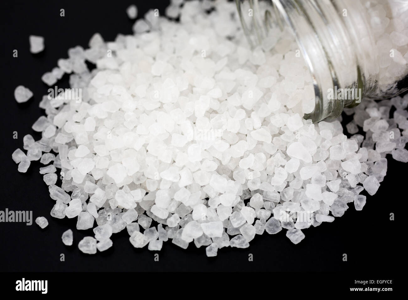 A glass container of sea salt spilling the large granules on to a black background. Stock Photo