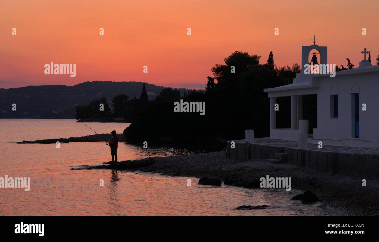 Fisherman at sunset next to Greek church by waterside at Porto Heli, Argolida, Peloponnese, Greece Stock Photo