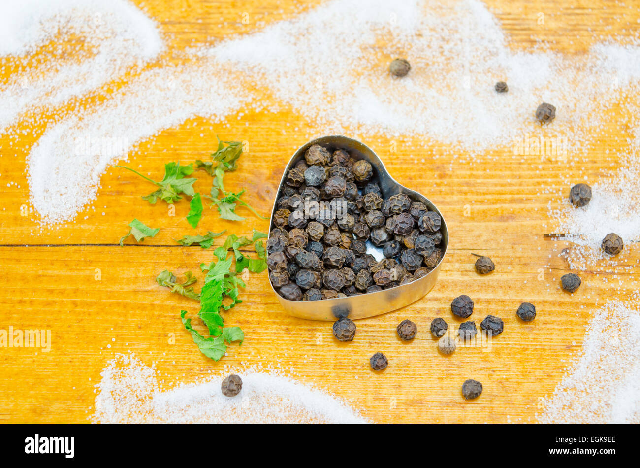 Black pepper in a hear shaped container decorated with basil on a wooden table sprayed with salt Stock Photo