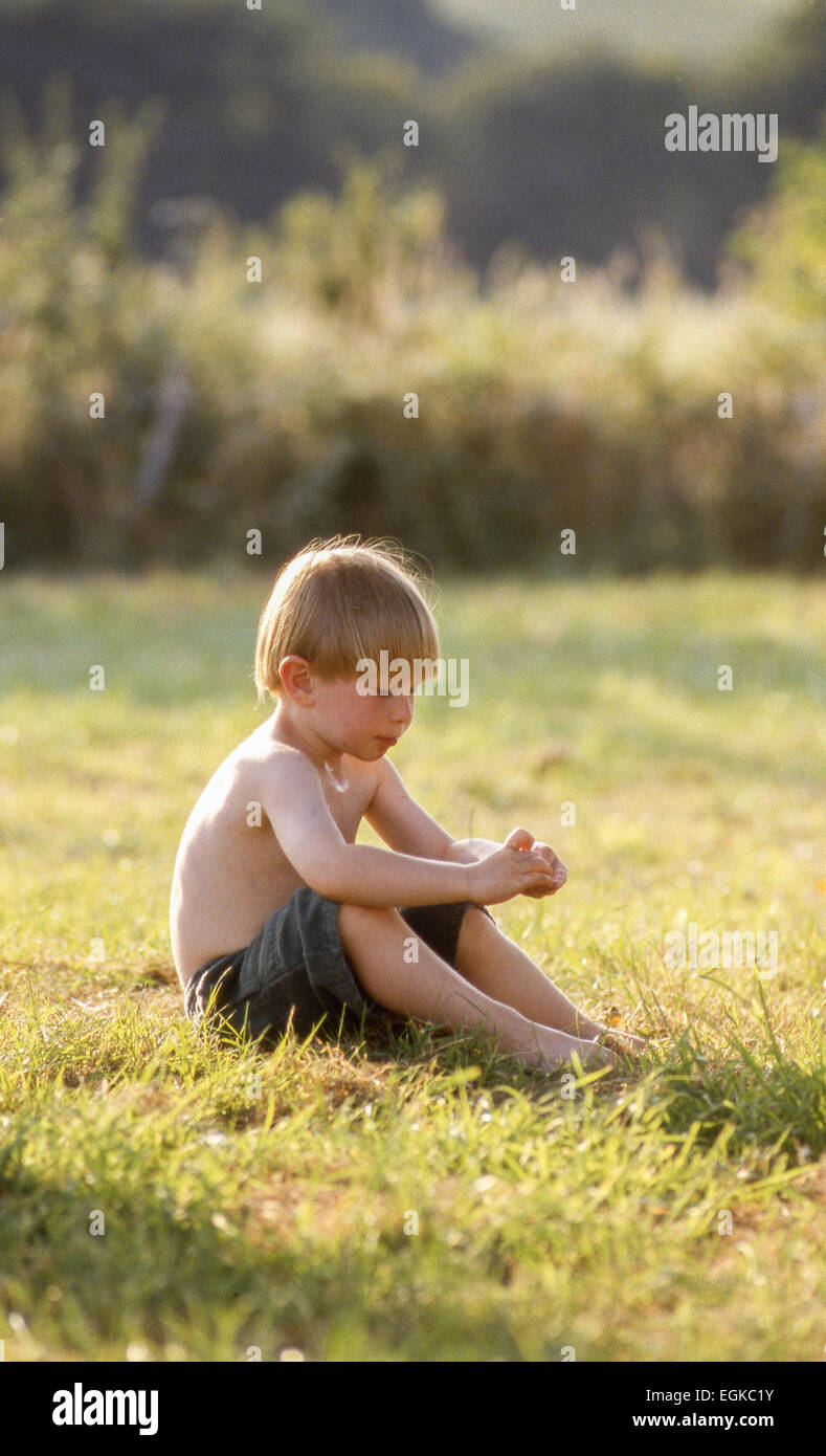 Young thoughtful barefoot boy sitting alone in sunny natural country green grass field with hedge in background Stock Photo