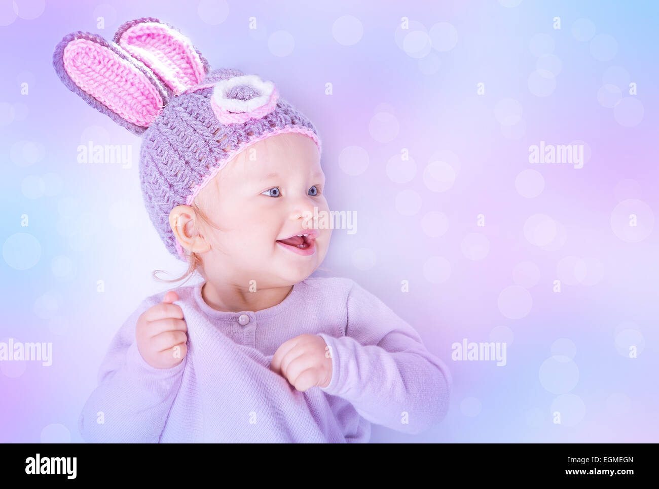 Portrait of happy cute baby girl wearing knitted hat with rabbit ears on purple blur background, beautiful Easter bunny Stock Photo