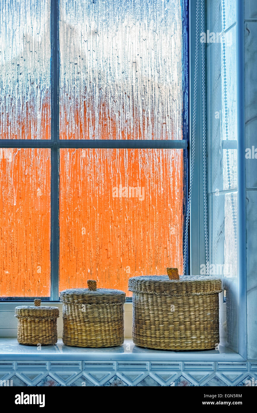 Set of three small wicker baskets sitting on a bathroom windowsill against a frosted window, with distorted orange exterior. Stock Photo