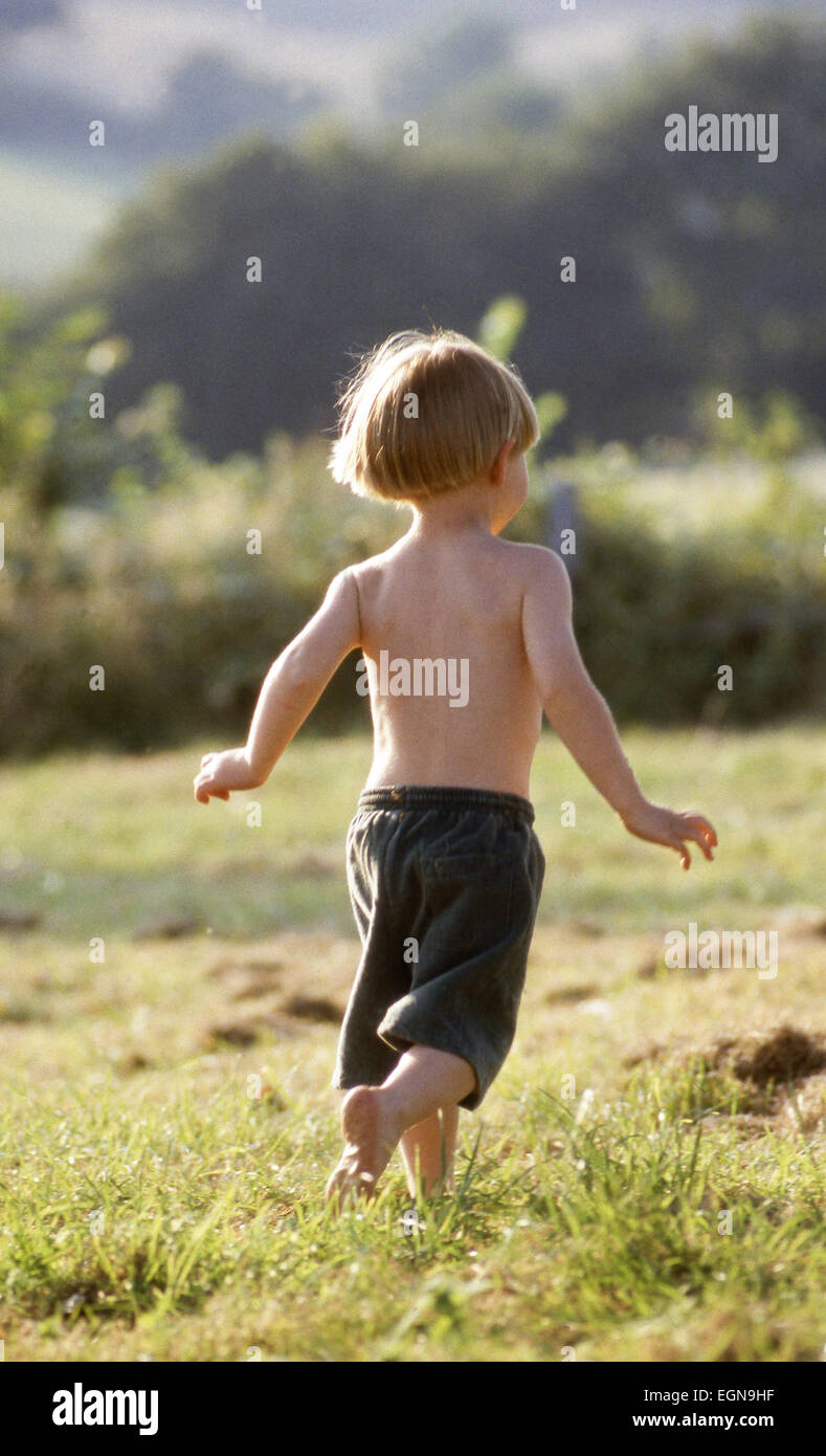 Young active happy barefoot boy running away in sunny natural country green field with trees and hedge in background Stock Photo