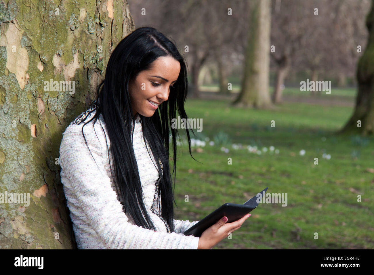 An attractive young woman reads an ereader under a tree Stock Photo