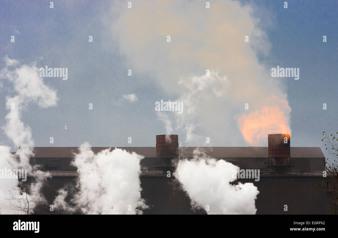 industrial pollution and flames pouring from roof top chimneys of grimy dirty steel mill with heat haze smoke and steam clouds Stock Photo