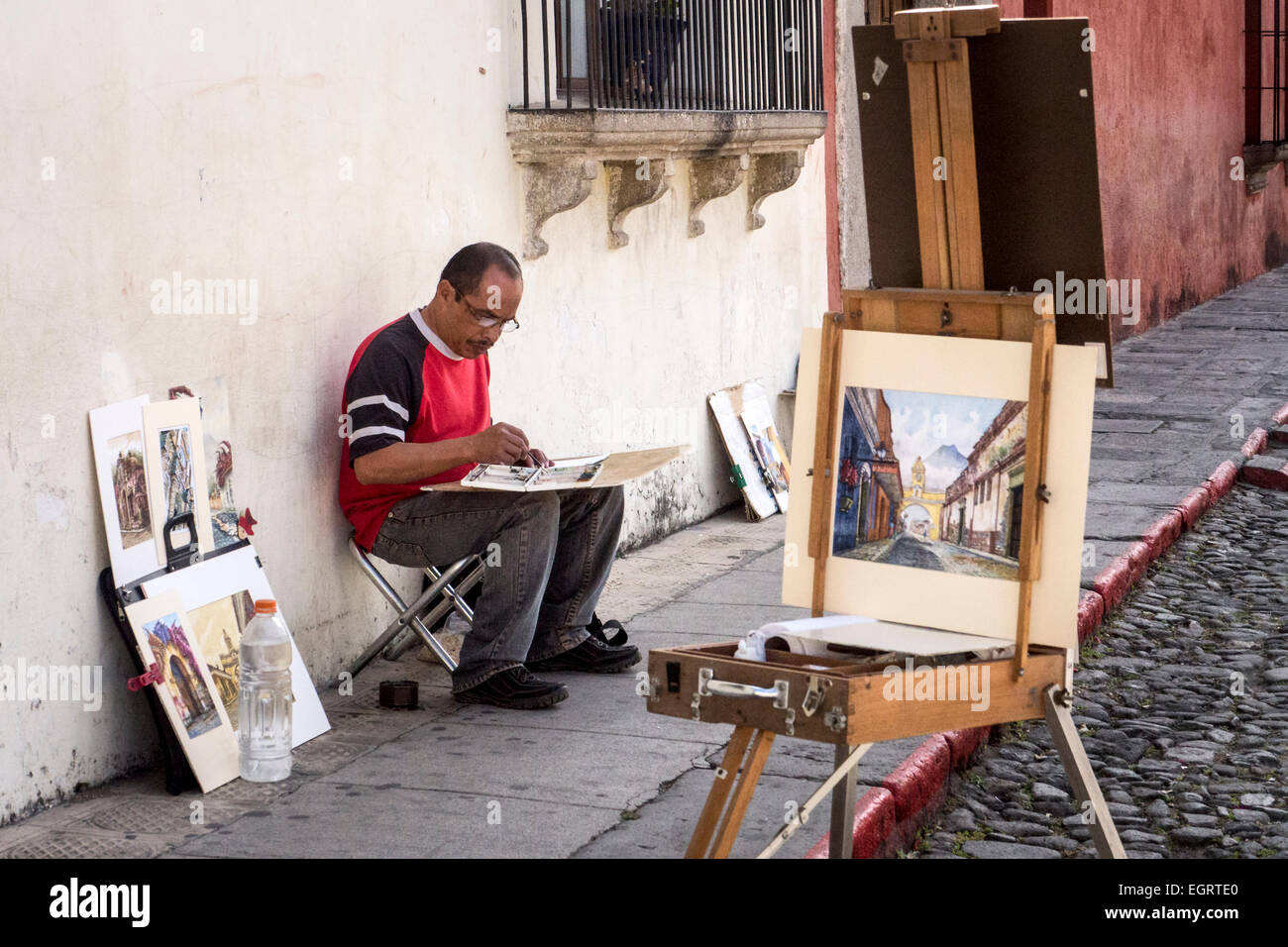 A n artist exhibiting his paintings for sale sits on a pavement in old Antigua and paints Stock Photo
