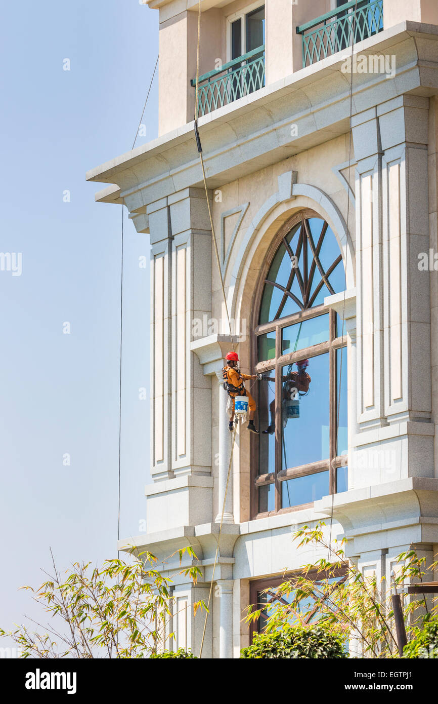 Window cleaner wearing orange overalls and red helmet, suspended from harness and ropes cleaning windows in Chennai, India Stock Photo