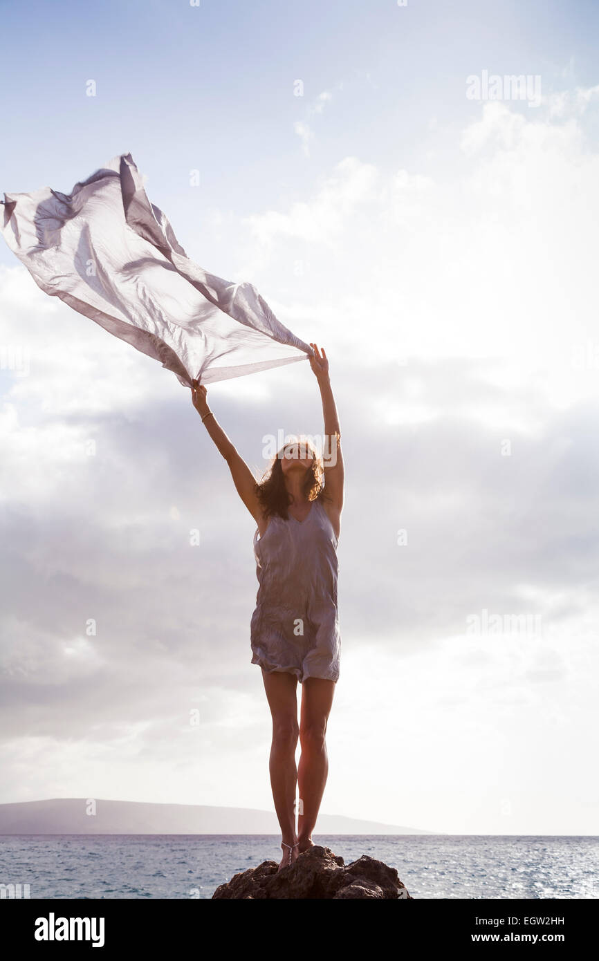 Woman standing on rock near ocean, holding fabric in the wind. Stock Photo