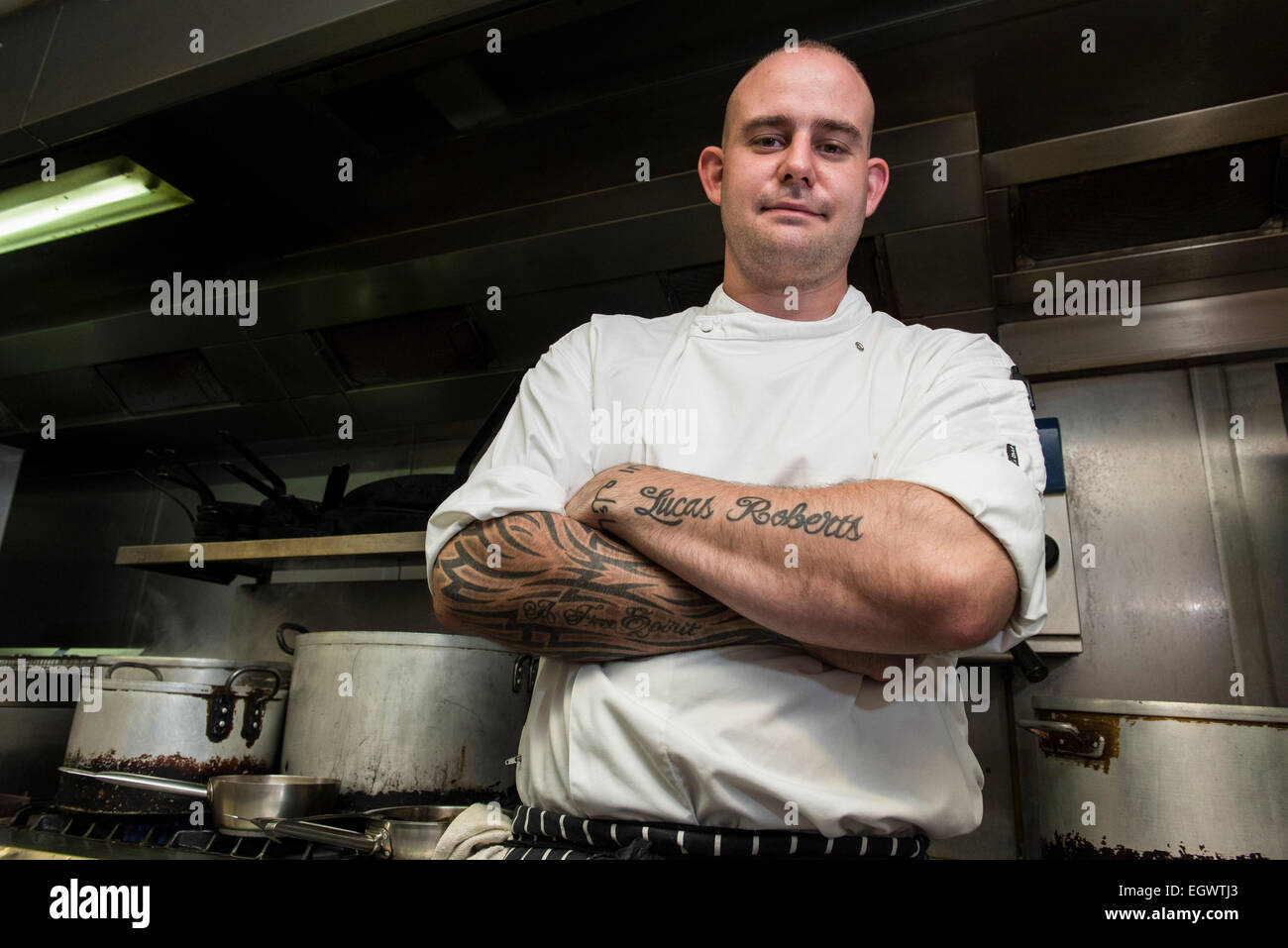 A professional mean ^ moody looking chef stands in his kitchen, looking annoyed with lots of cooking equipment in the background Stock Photo