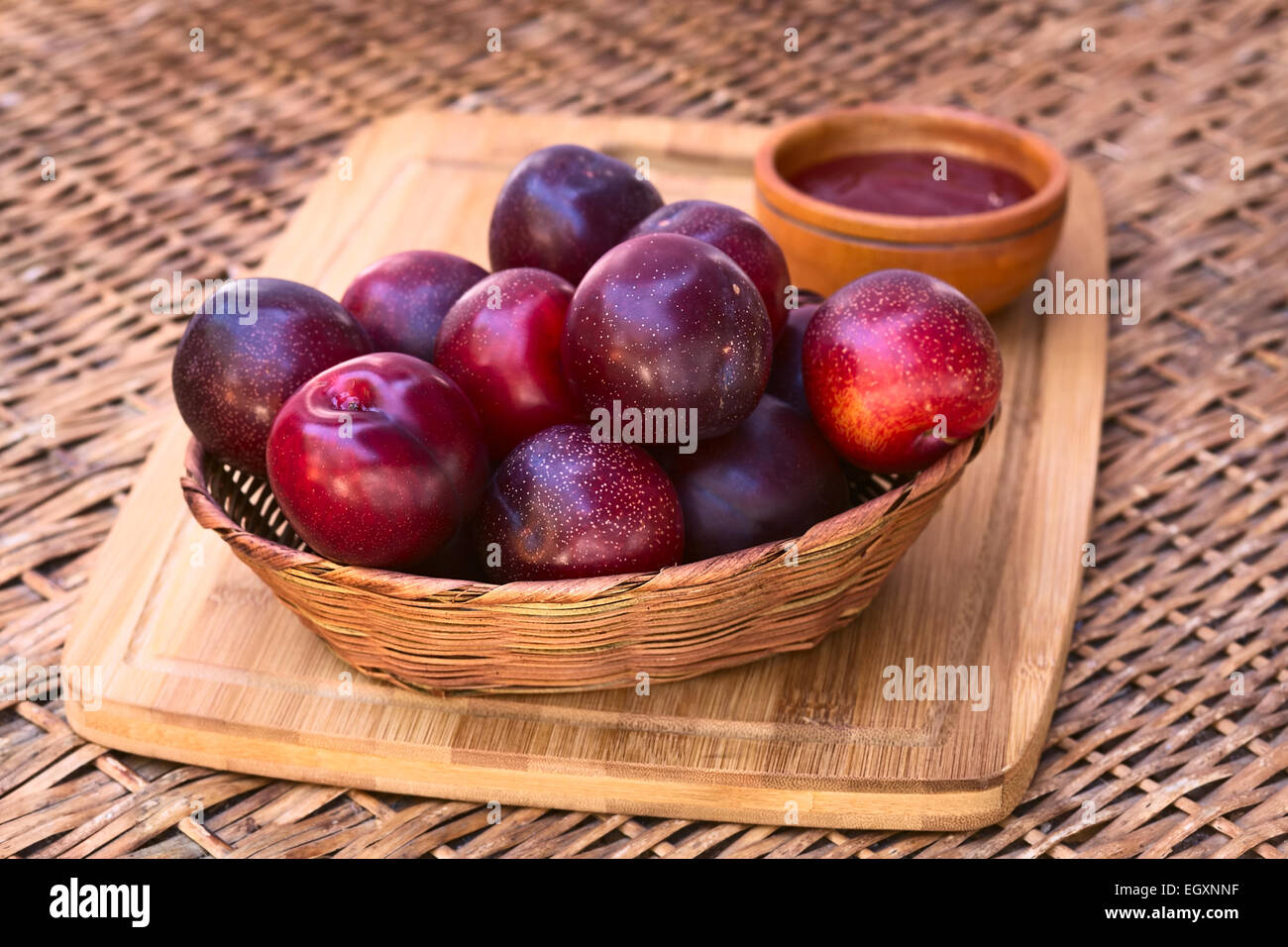 Woven basket filled with satsuma plum with small bowl of plum jam in the back on wooden board photographed with natural light Stock Photo