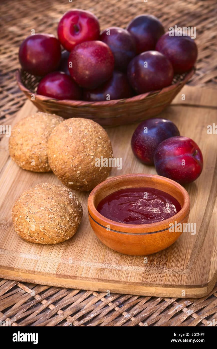 Small wooden bowl of plum jam with wholegrain buns and satsuma plums on wooden board photographed with natural light Stock Photo