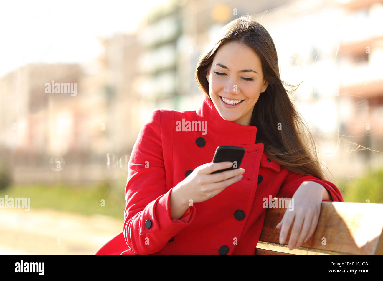 Girl texting on the smart phone sitting in a park wearing a red jacket and sitting in a bench in a park Stock Photo