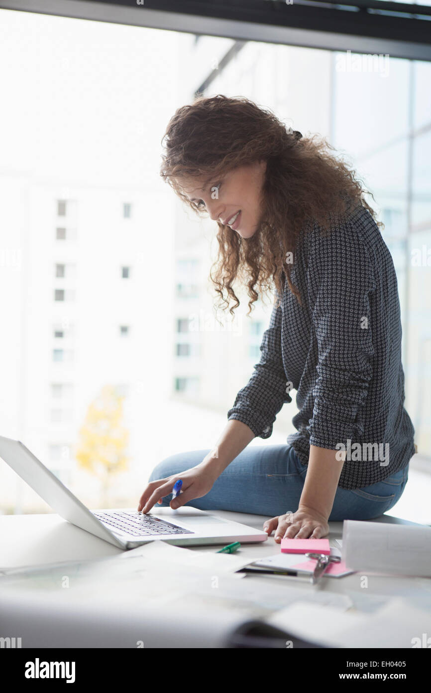 Young woman sitting on desk in office using laptop Stock Photo