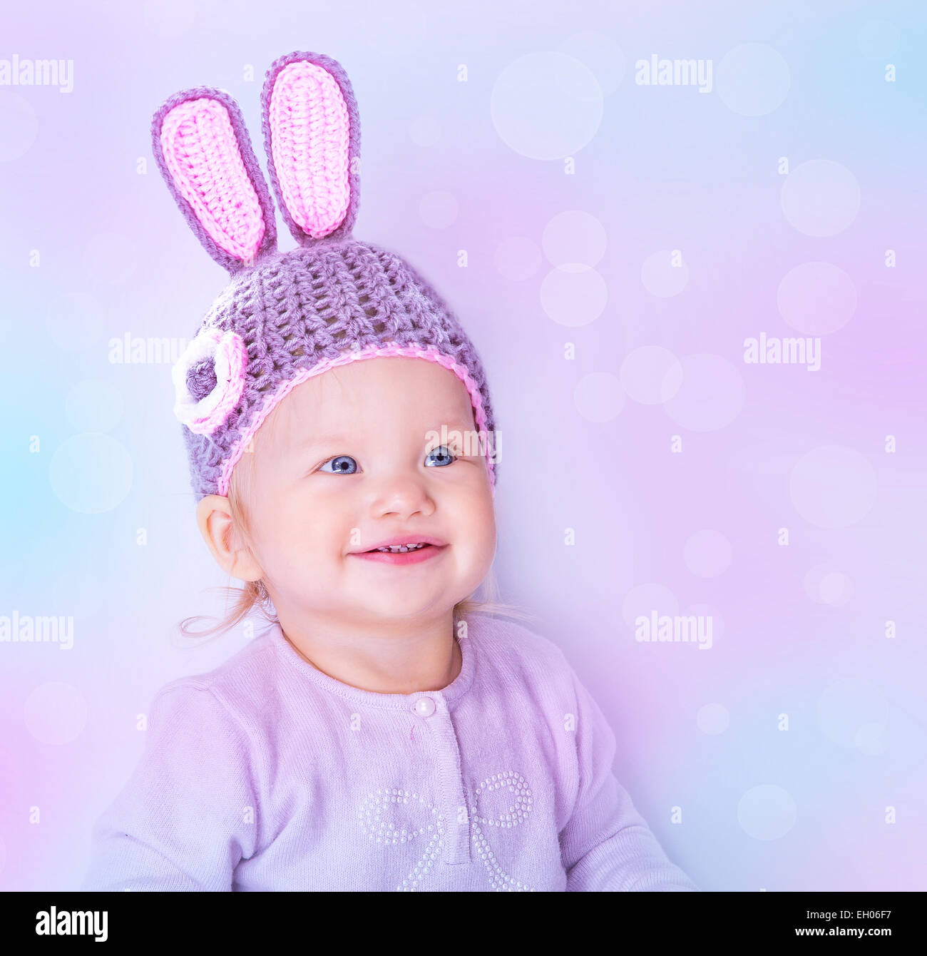 Portrait of happy cute baby girl wearing knitted hat with rabbit ears on purple blur background, beautiful Easter bunny Stock Photo
