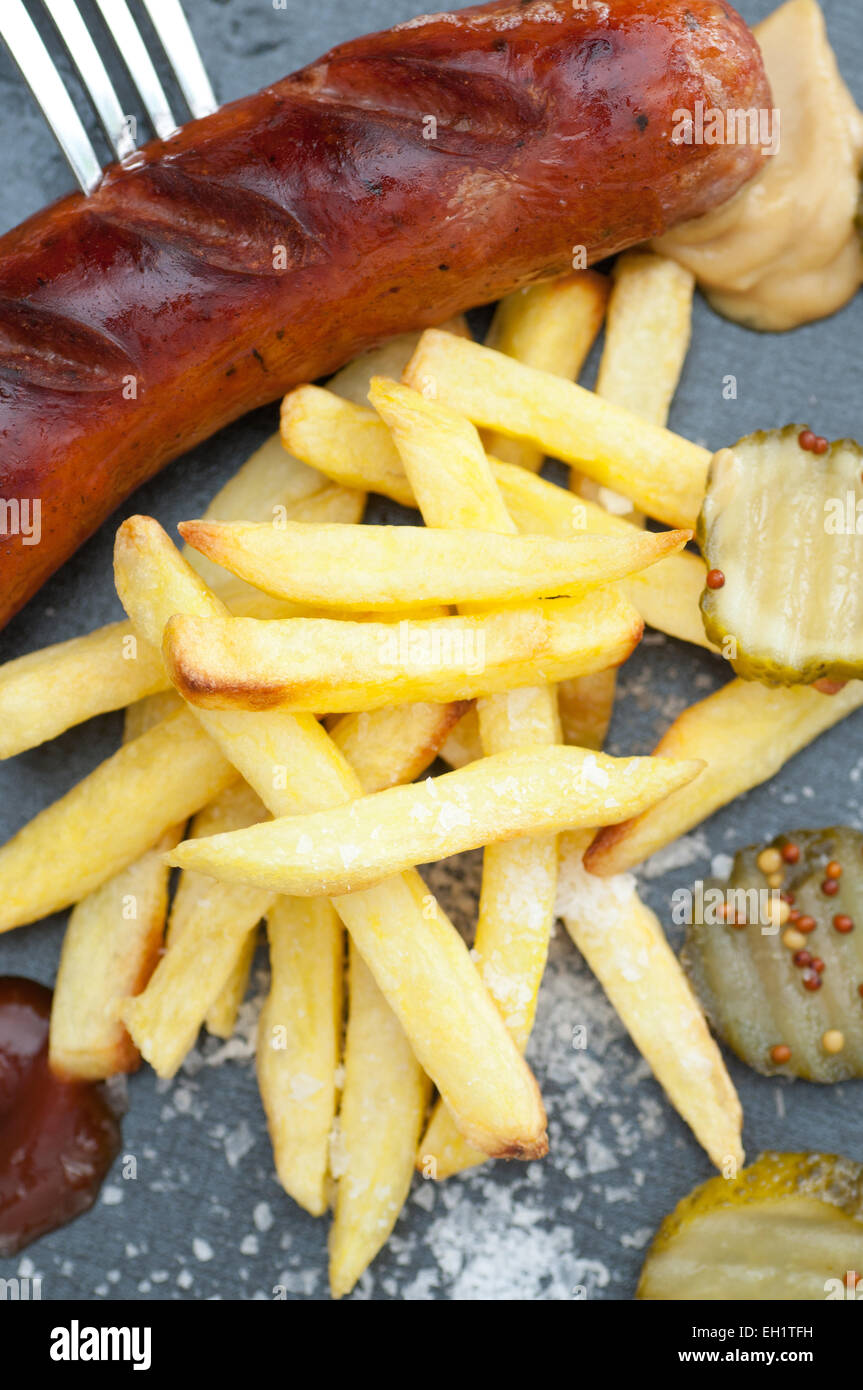 Grilled sausage with french fries, pickled cucumber, mustard, ketchup and sea salt. Stock Photo