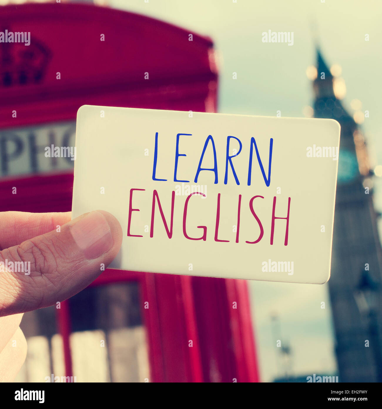 the hand of a man holding a signboard with the text learn english written in it with a red telephone booth and the Big Ben in th Stock Photo