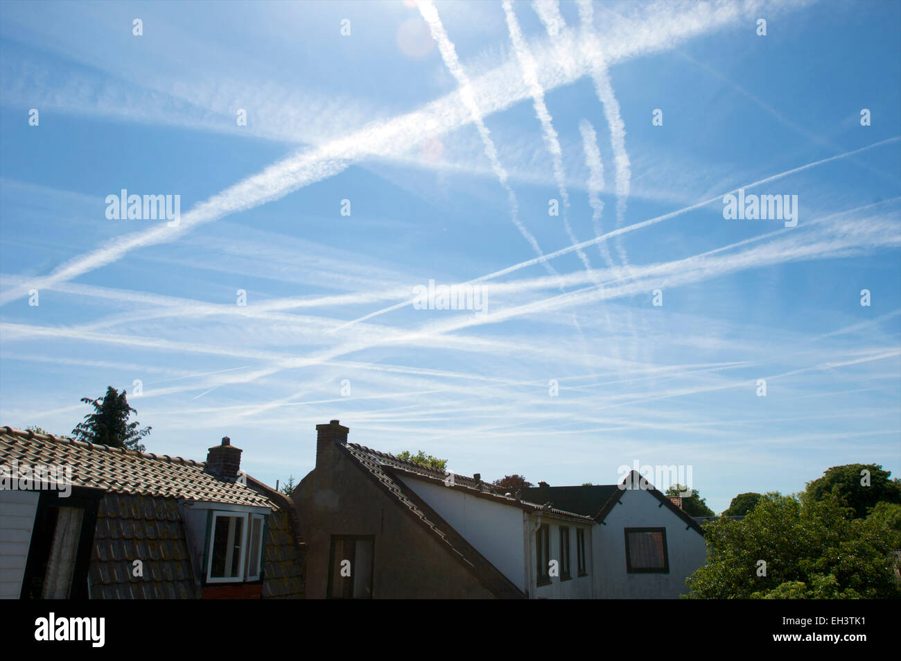 Chemtrails and contrails in a blue sky above houses Stock Photo