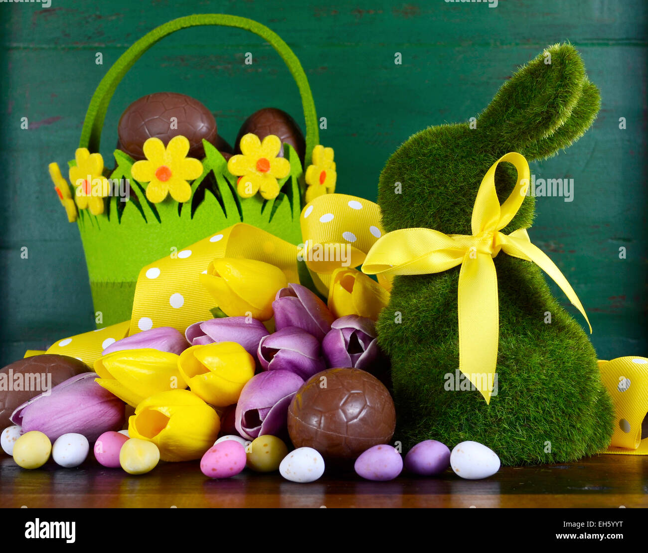 Happy Easter green moss grass bunny rabbit with basket of chocolate eggs and spring tulips against a green wood background. Stock Photo