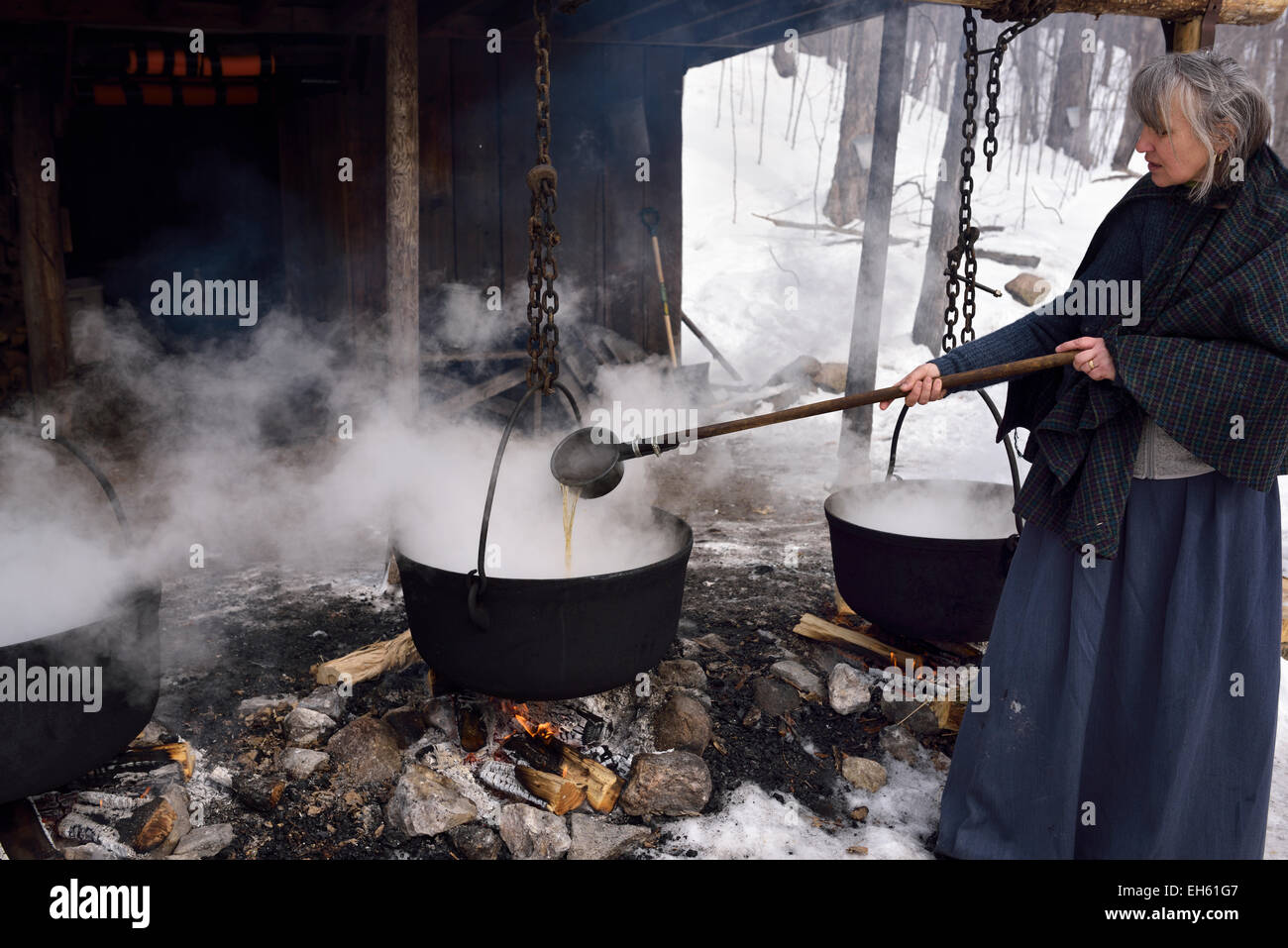 Woman in pioneer clothes ladling evaporated sap in steaming cast iron pots outdoors in winter to produce maple syrup Ontario Canada Stock Photo