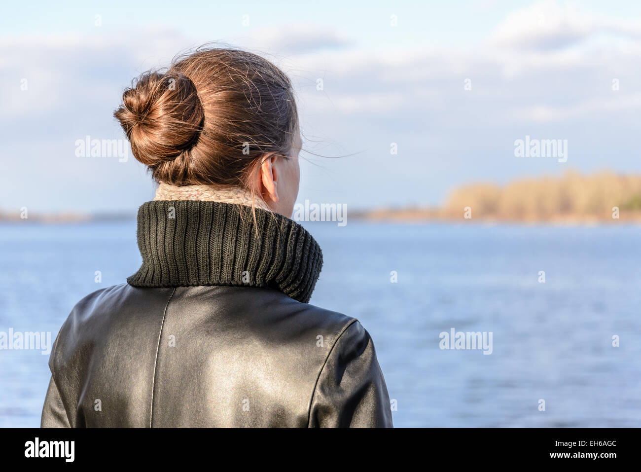 A woman with a chignon and a black leather coat is watching the landscape close to the lake or the river during a sunny day with Stock Photo
