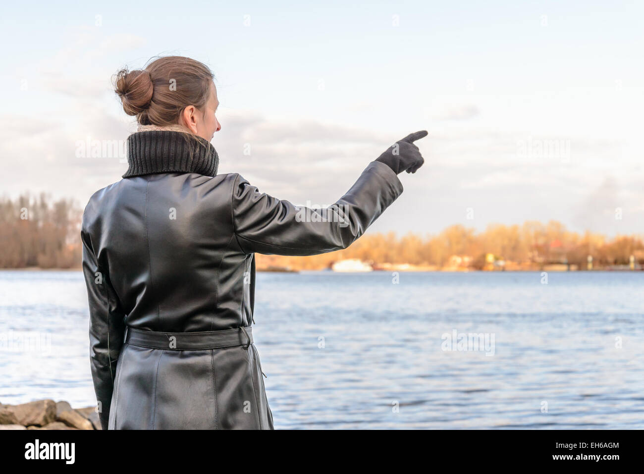 An adult with a chignon and wearing a black leather coat stays close to the river and point her finger to indicate something int Stock Photo