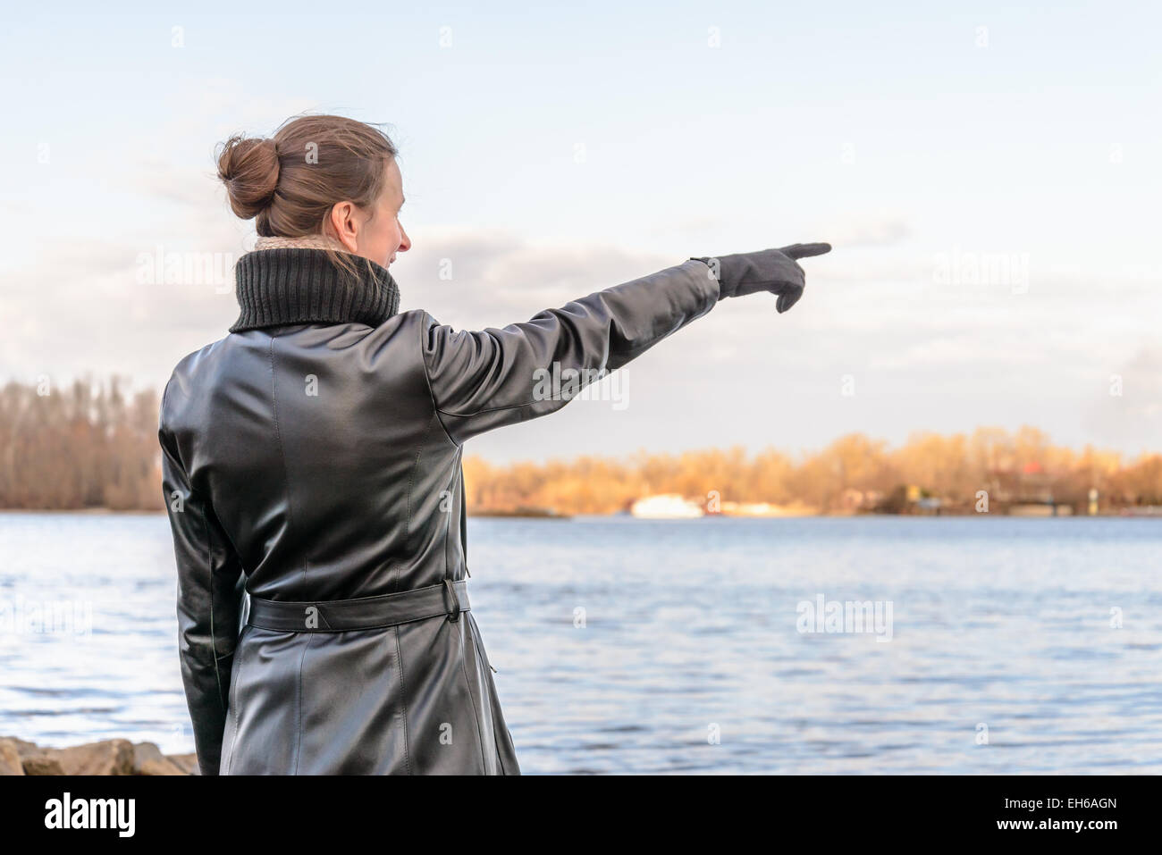 An adult with a chignon and wearing a black leather coat stays close to the river and point her finger to indicate something int Stock Photo