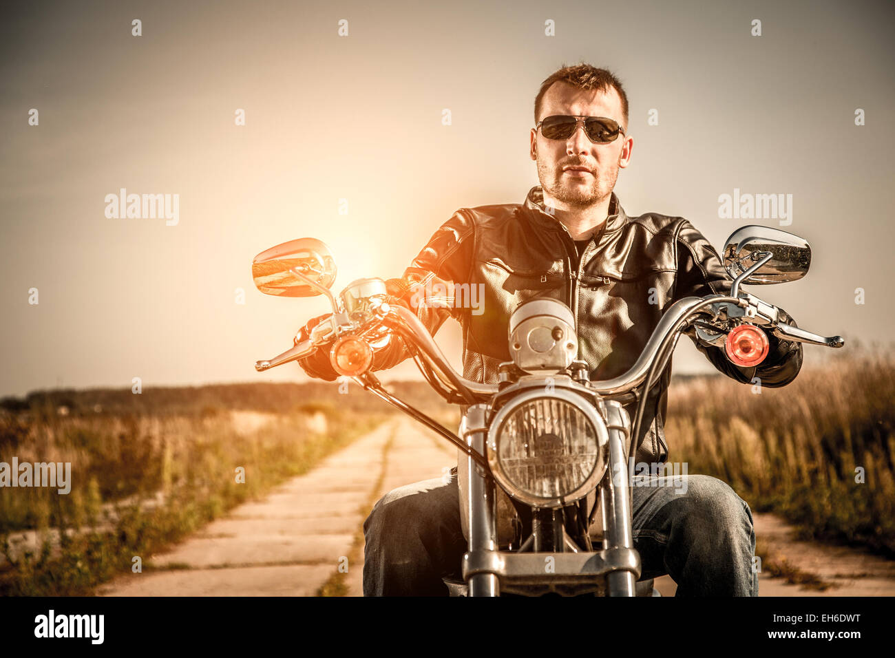 Biker man wearing a leather jacket and sunglasses sitting on his motorcycle looking at the sunset. Stock Photo