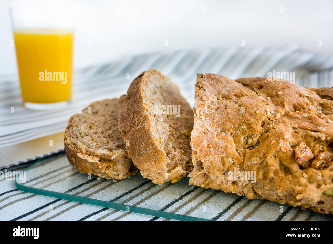 A close-up of a loaf of wholesome bread and bread knife with a glass of orange juice on a striped cloth and glass cutting board Stock Photo