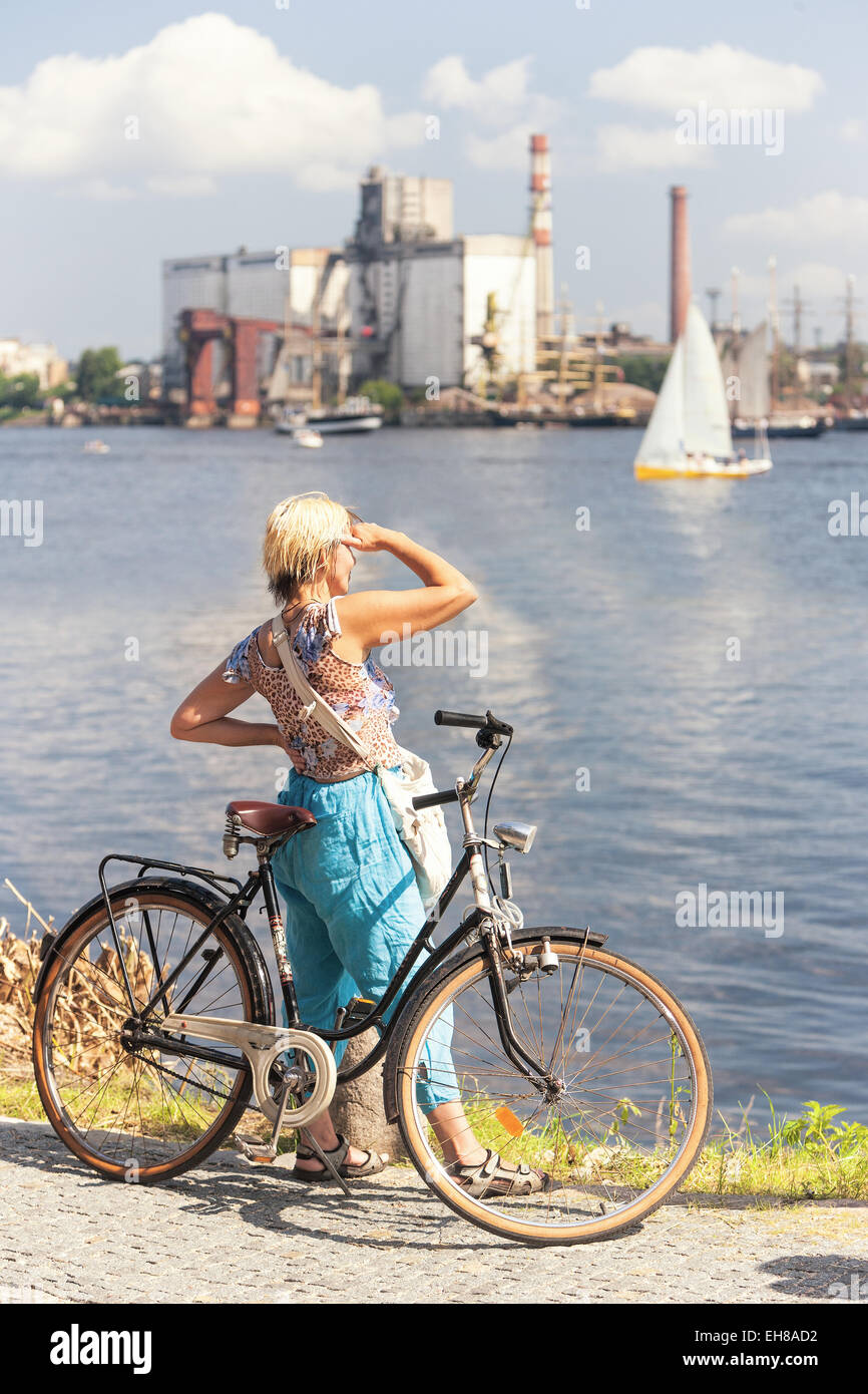 Girl with bicycle looking at the boat and the city by the river shielding her eyes from the sun. Riga, Latvia Stock Photo
