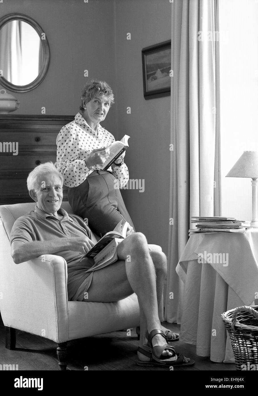Retirement age couple in a front room positioned to a corner by the side of a window. The man is seated in a comfortable armchair, the woman standing and resting on the back of the chair by his side. The couple are holding books and looking into camera. They are relaxed and smiling. The man is wearing shorts and sandals, the woman trousers and long sleeved patterned blouse. The room setting is traditional, with curtain, a painting and mirror on the wall, a cloth covered occasional table and a wicker basket (partly cropped out of the shot). The image is black and white and portrait format. Stock Photo