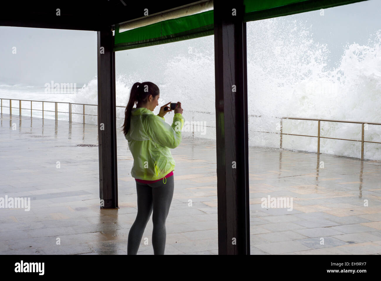 women photographing large crashing waves aginst broadwalk with mobile phone, dressed in running gear Stock Photo