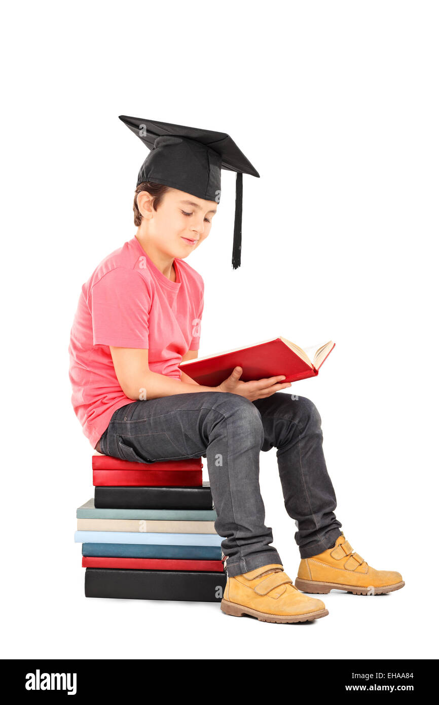 Kid with mortarboard reading a book seated on a stack of books isolated ...