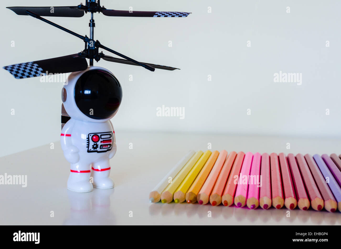 A child's toy spaceman looks towards a set of coloring crayons laid in a row on a white surface. There is some reflected detail. Stock Photo