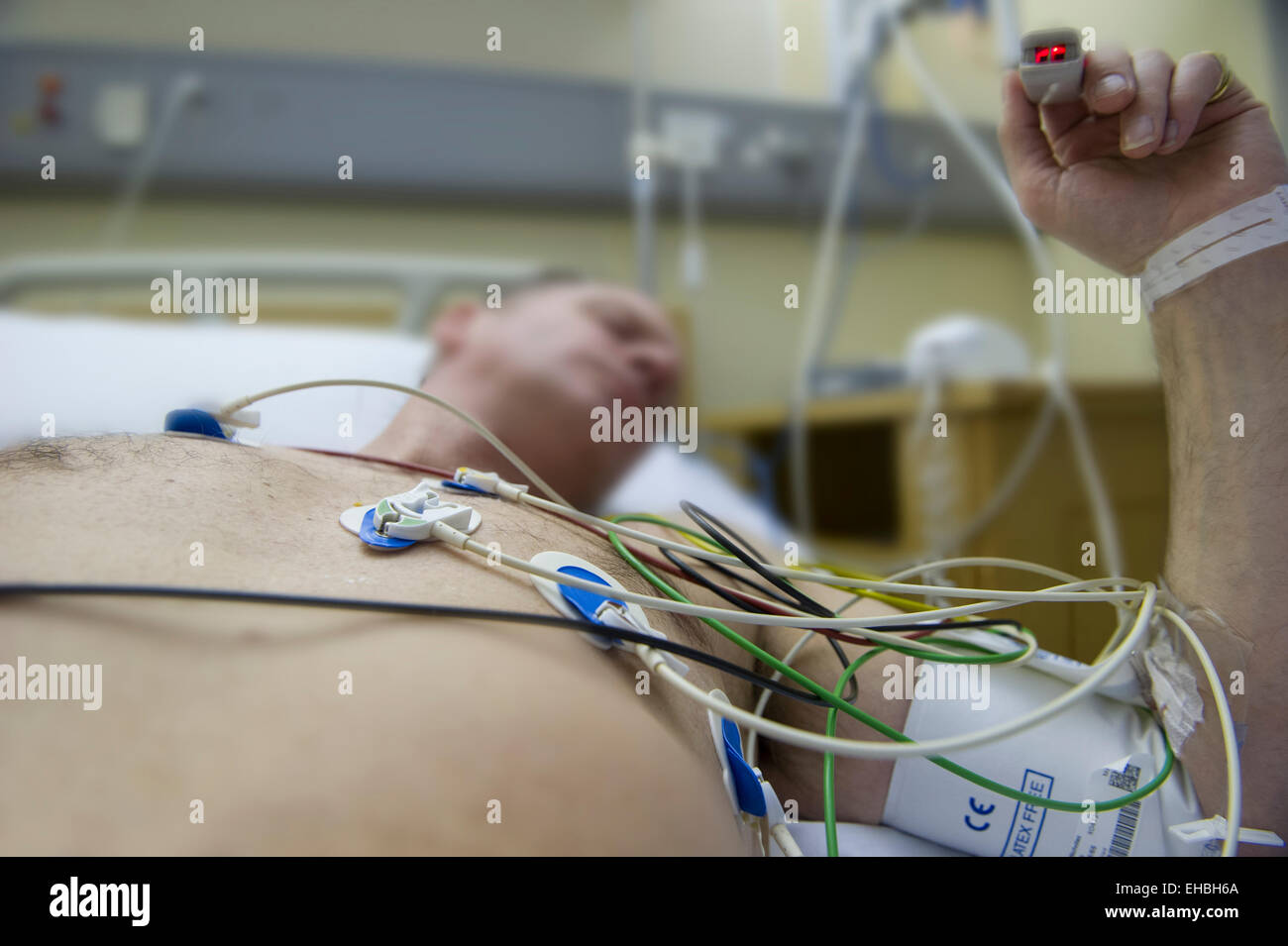 A man lies in a hospital bed with heart monitors to assess the risk of a heart attack, his pulse being taken on his finger Stock Photo