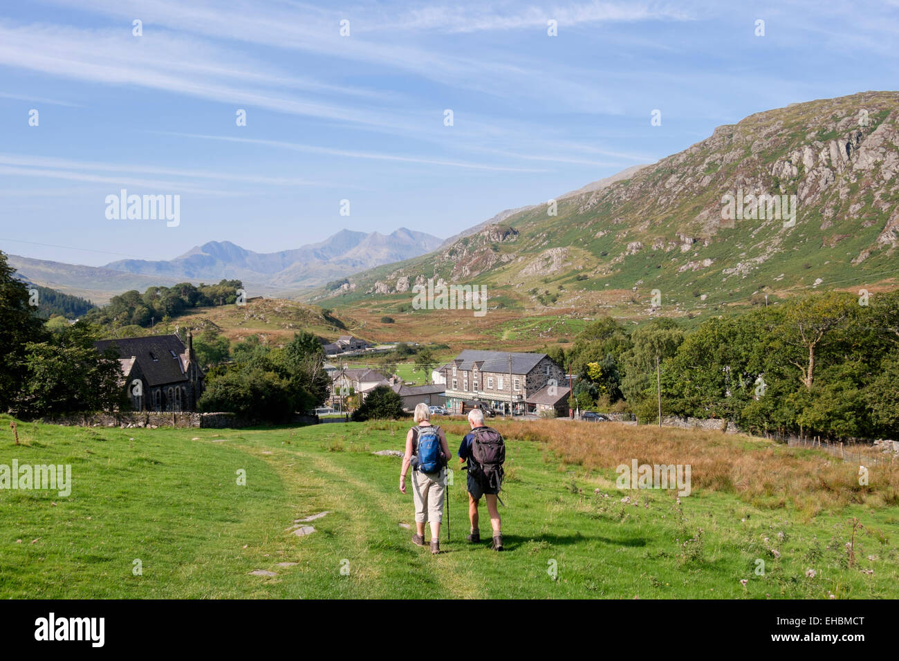 Two walkers walking down hillside to village with view to Snowdon in distance in Snowdonia National Park (Eryri). Capel Curig Conwy Wales UK Britain Stock Photo