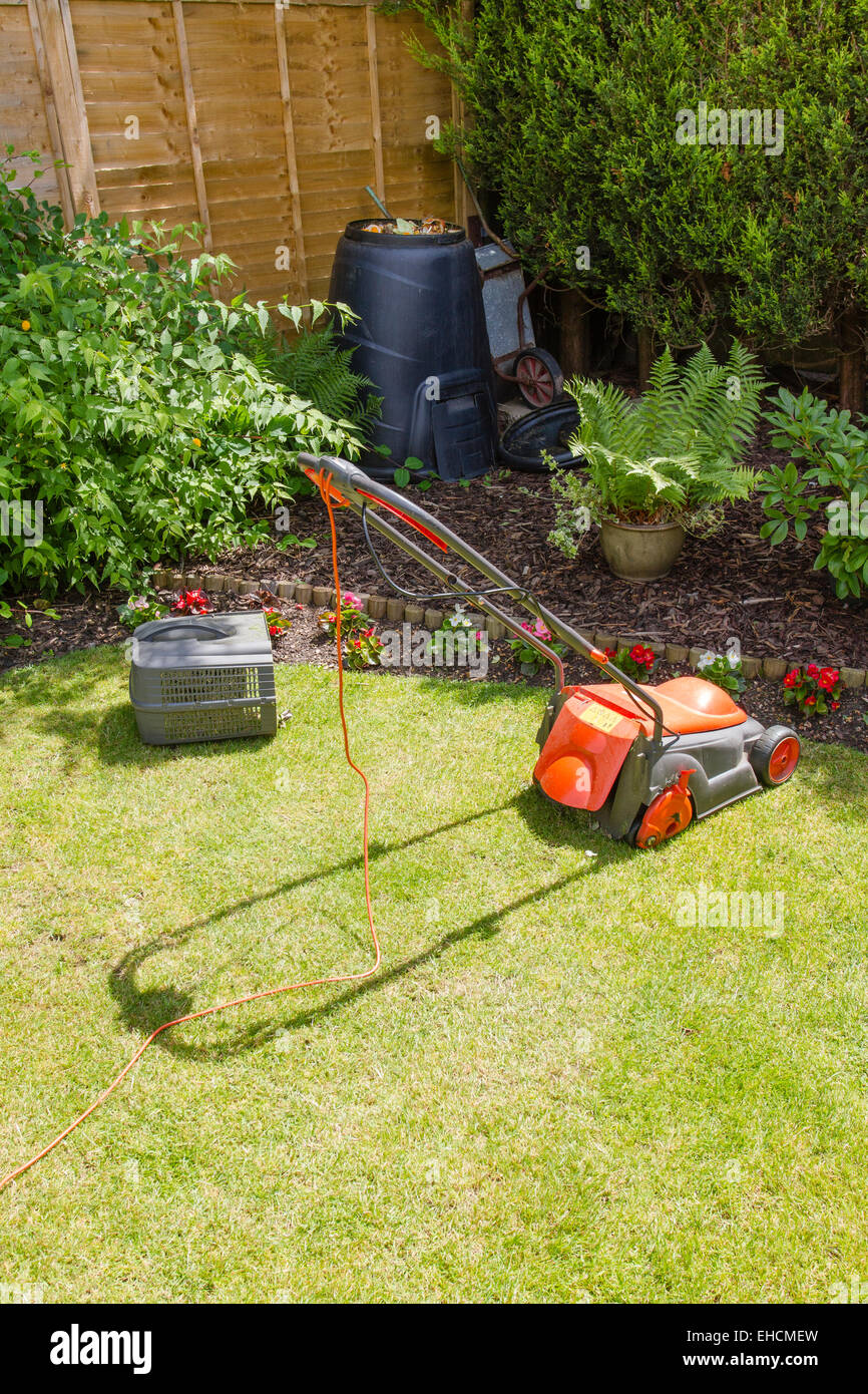 An electric lawn mower with grass box detatched on a lawn near to an open compost bin and a stored wheelbarrow Stock Photo
