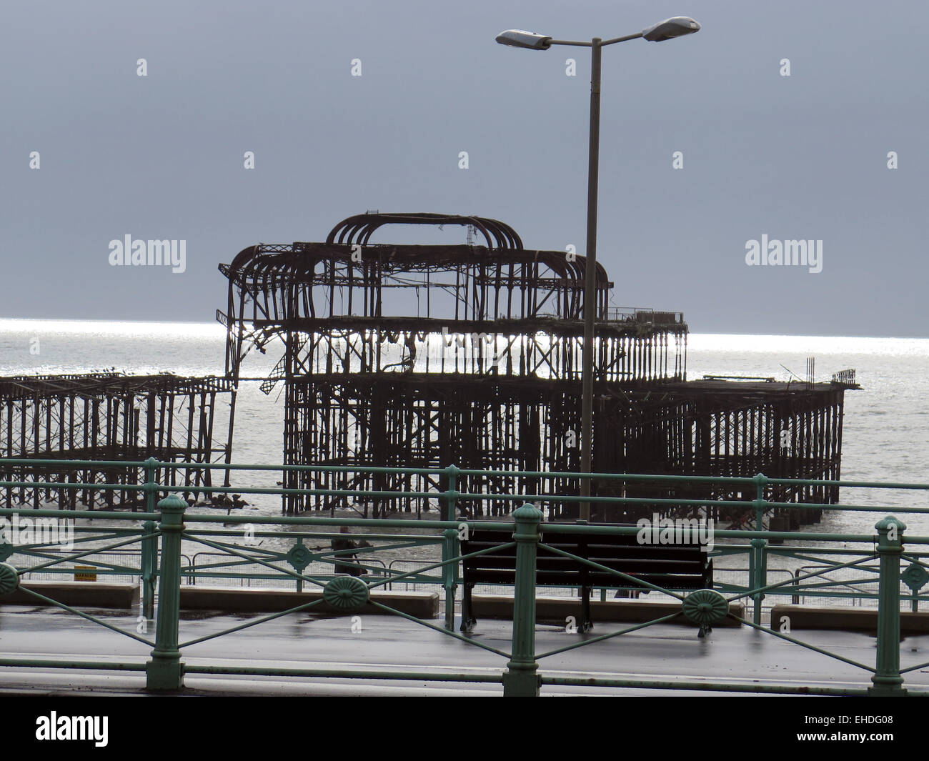 The Arson attacked ruin of the West Pier, Brighton designed by Eugenius Birch, seen across the promenade with decorative railings and benches to a glistening sea. Stock Photo